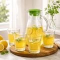 Glass water jug with green lid and four matching drinking glasses on a dining table.